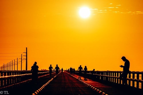 Where to watch the sunset in marathon fl | 7 stunning spots 4 Silhouettes of people walking and standing on a long bridge at sunset, with a bright orange sky and the sun shining above. Some people are looking at their phones, and railings line both sides of the bridge.