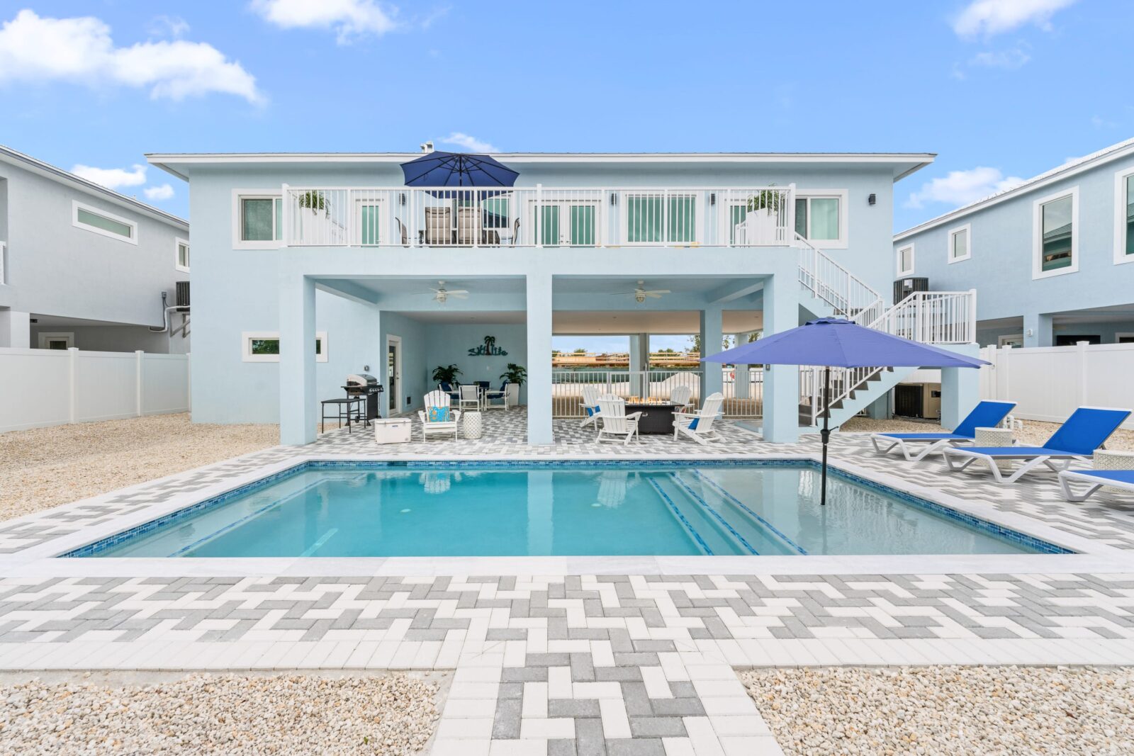 A modern blue pearl marathon fl light blue two-story house with a large outdoor swimming pool, lounge chairs under blue umbrellas, a balcony, and a patio area with seating and a grill—surrounded by a paved area and white fence.