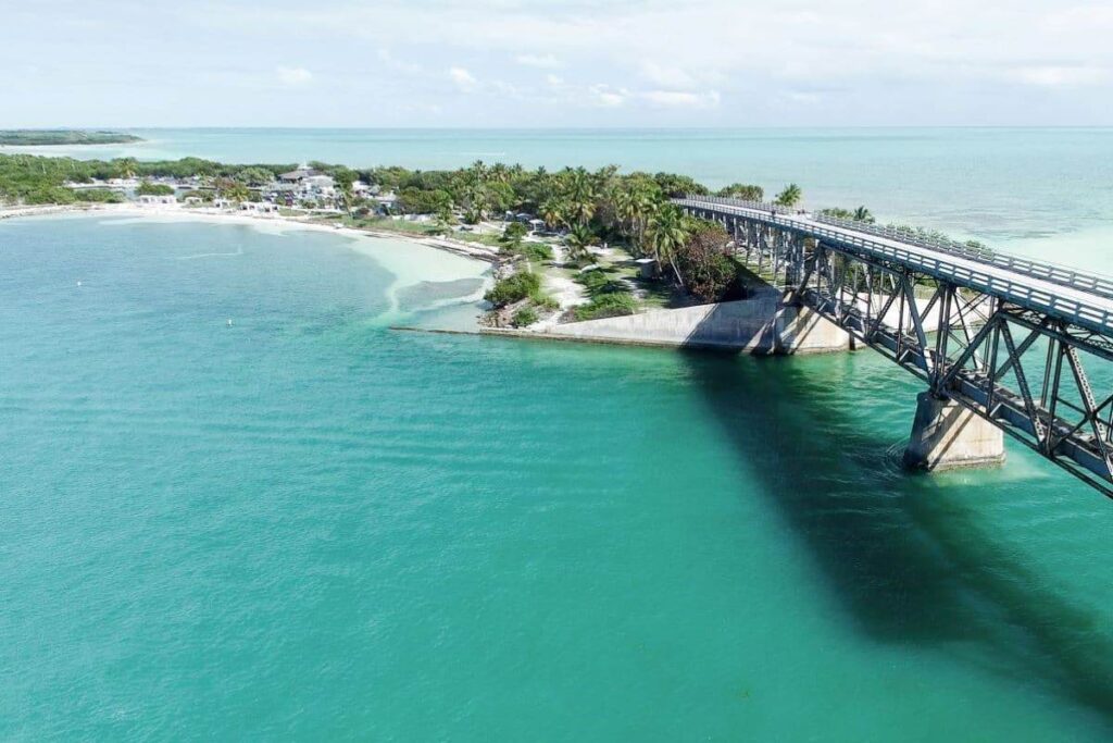 A long bridge stretches over turquoise water toward a tropical shoreline in the florida keys, where palm trees, sandy beaches, and scattered buildings await under a partly cloudy sky—perfect for spring break adventures.
