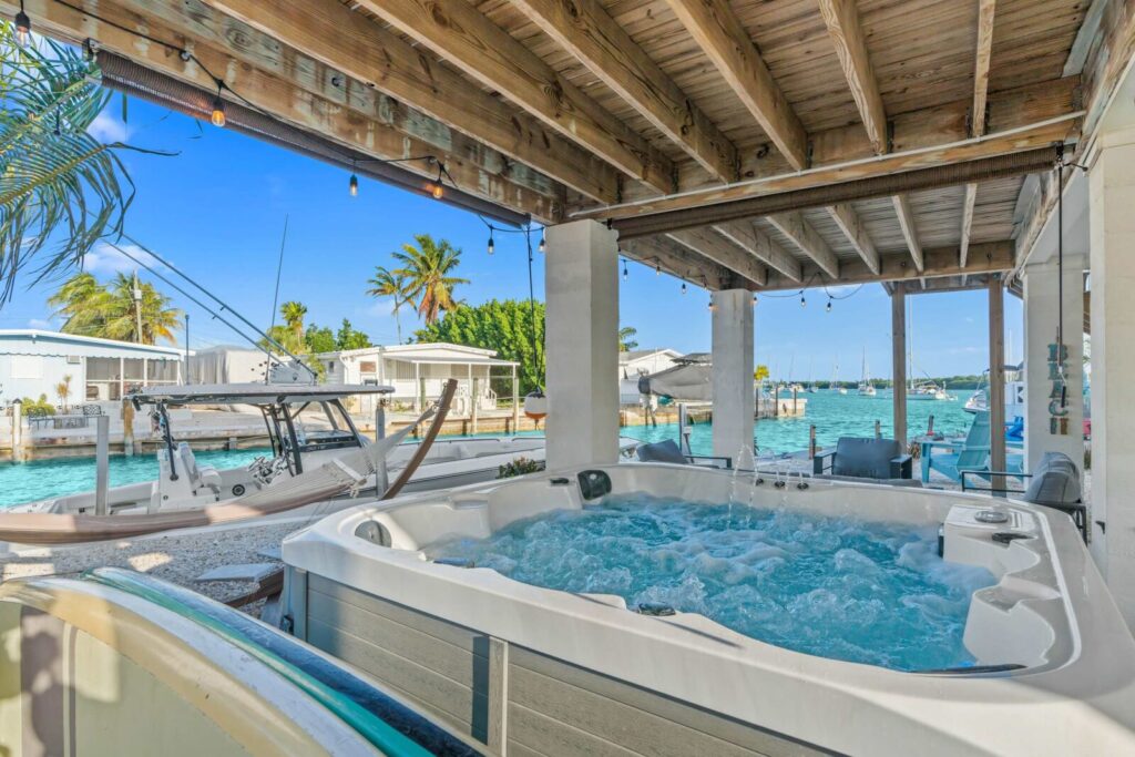 A bubbling hot tub sits under a wooden patio roof beside a turquoise canal, with boats, palm trees, and tropical homes in the background on a sunny day.