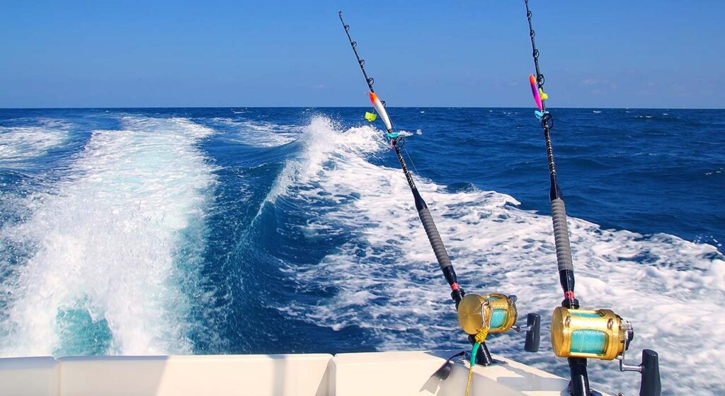 Two fishing rods are mounted on the back of a boat moving swiftly through deep blue ocean water, leaving a white foamy wake behind under a clear blue sky.