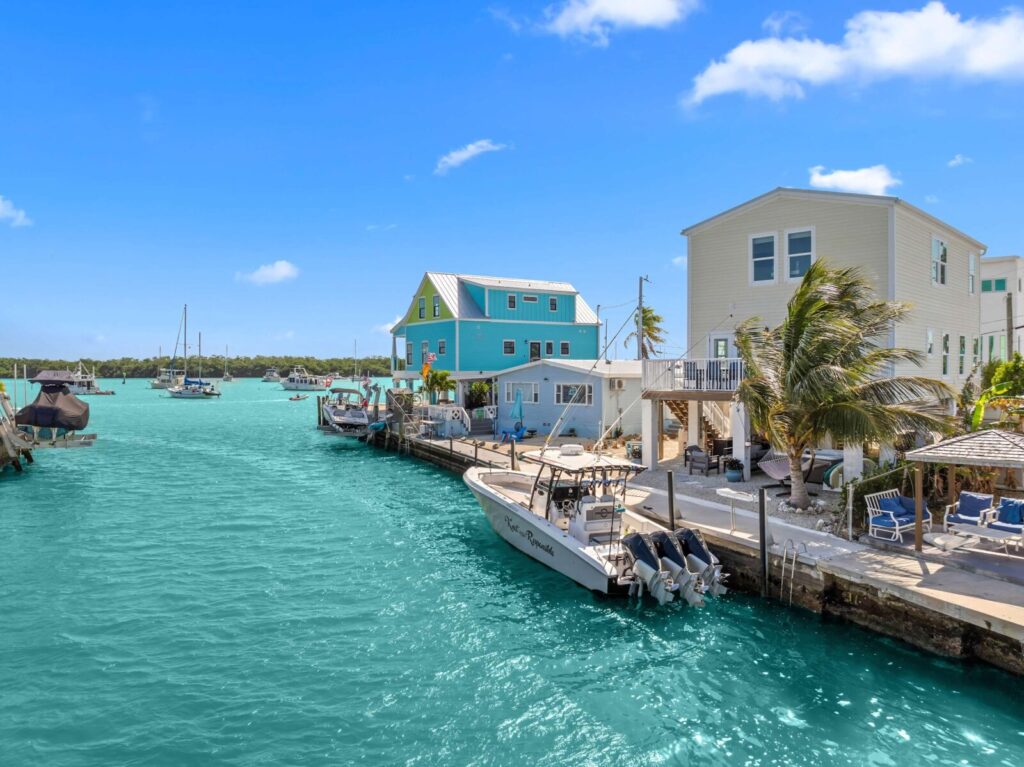 Turquoise water lined with boats and colorful waterfront houses under a bright blue sky. Palm trees and outdoor seating areas are visible, creating a relaxed, tropical atmosphere. Waterfront vacation rental in florida keys | mermaid manor