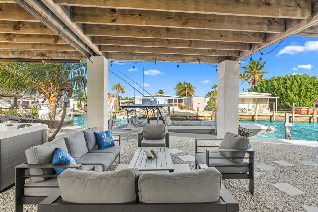 Outdoor seating area under a wooden pergola with cushioned sofas and chairs, overlooking a dock and canal with boats, palm trees, and clear blue sky in the background.