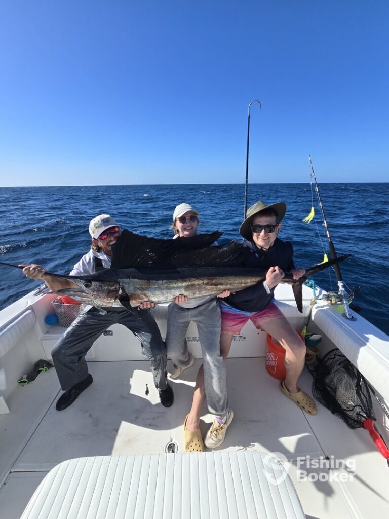 Three people on a boat hold a large sailfish together, smiling at the camera. The ocean is calm and blue, with fishing rods visible in the background. The fishing booker logo is in the lower right corner.