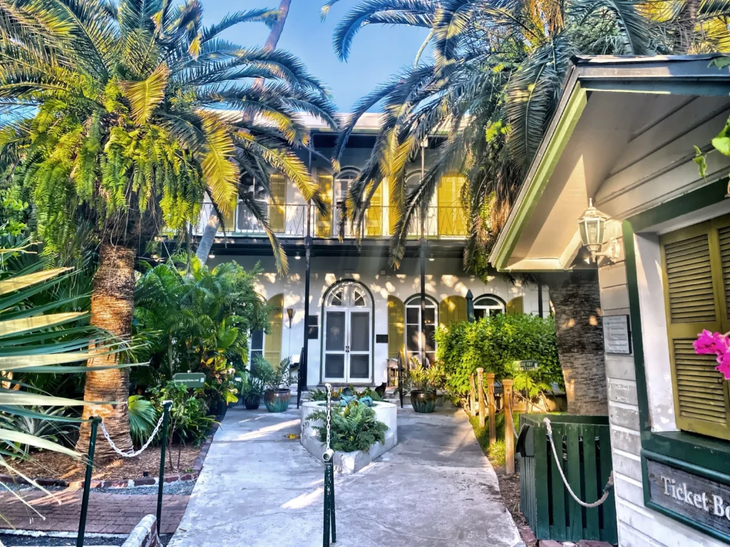 A two-story house with white doors and large windows is surrounded by lush palm trees and greenery, perfect for a spring break in the florida keys. Sunlight filters through the trees, casting dappled shadows on the walkway to the entrance.
