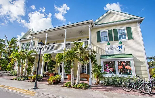 A two-story, light-yellow building with green shutters, palm trees out front, a porch with white columns, and a sign reading the key west butterfly beside bikes—a perfect spring break in the florida keys scene.