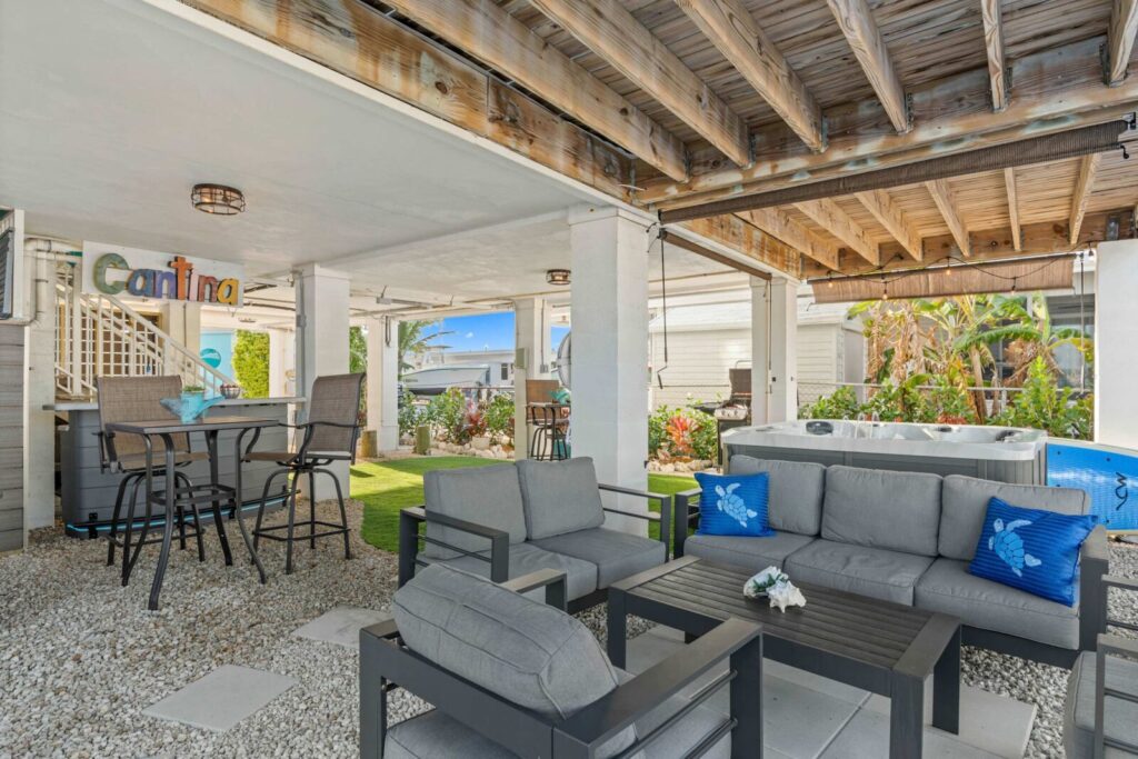 Outdoor covered patio with gray cushioned seating, a black coffee table, small bar area labeled cantina, and a hot tub. Pebble flooring, bright daylight, and tropical plants in the background create a relaxing atmosphere.