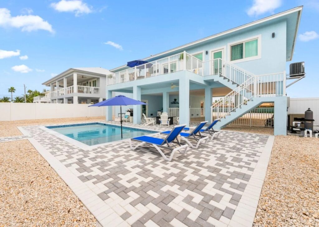 A modern two-story light blue house, known as blue pearl marathon fl, features an outdoor pool, poolside lounge chairs under a blue umbrella, a patterned stone patio, and a white staircase leading to an upper deck under clear sunny skies.