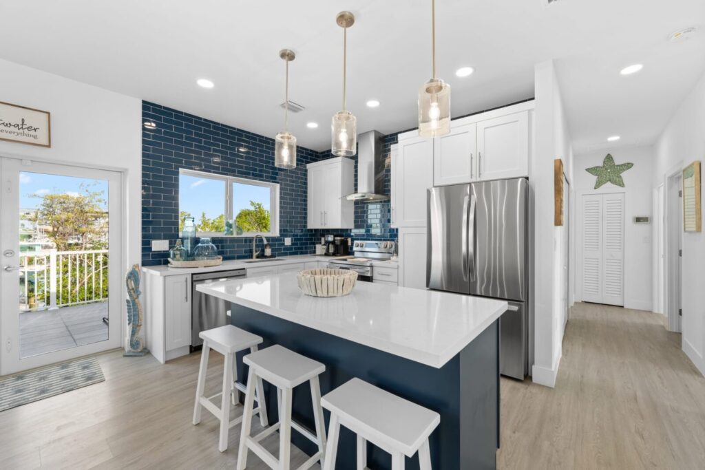 Modern kitchen with white cabinets, a navy island with white countertop, three bar stools, stainless steel appliances, blue subway tile backsplash, pendant lights, and a window showing greenery outside.