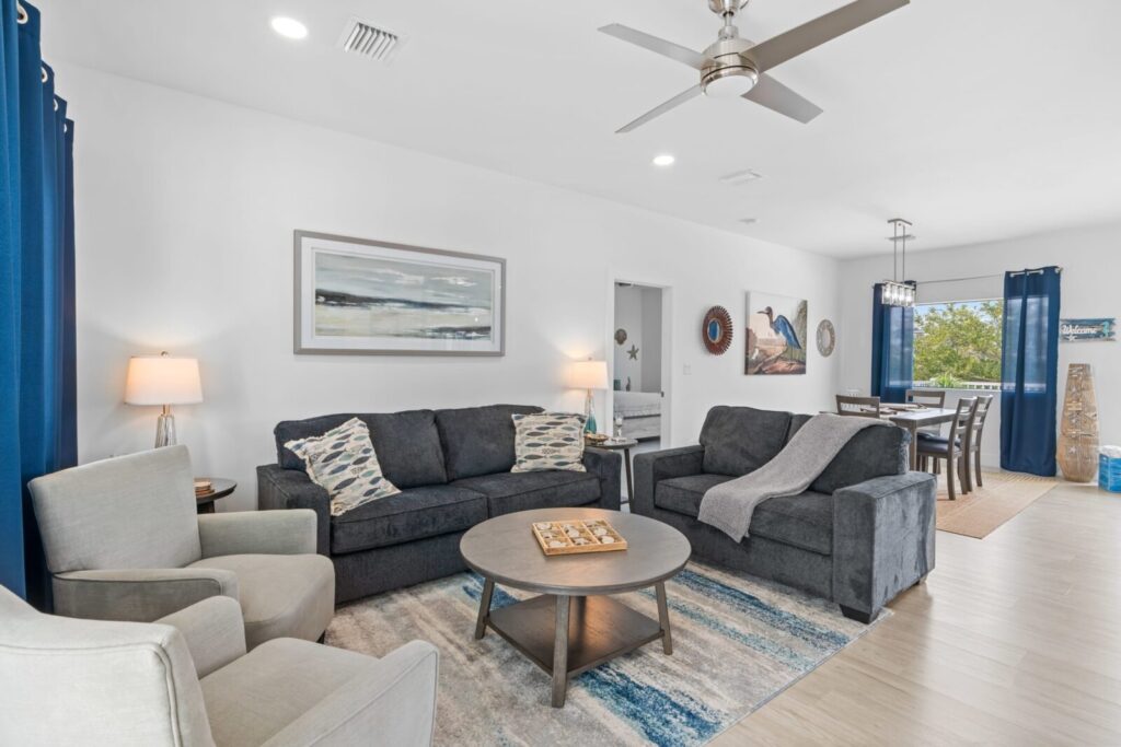 A modern living room with gray sofas, two gray armchairs, a round coffee table, blue curtains, coastal artwork, and a dining area visible in the background. The space is bright with white walls and wood floors.