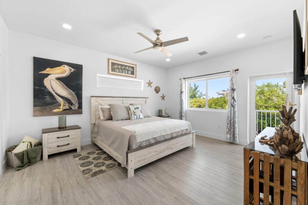 Bright bedroom with a large bed, pelican artwork above the nightstand, light wood furniture, ceiling fan, windows with curtains, and a door leading to a balcony with trees visible outside.