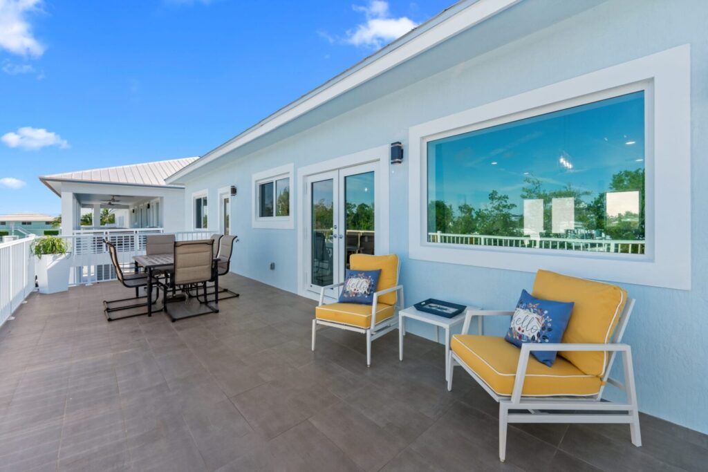 A spacious outdoor patio with yellow-cushioned chairs, a small table, and a dining set. The area has large windows, a light blue wall, and overlooks greenery under a bright blue sky with a few clouds.