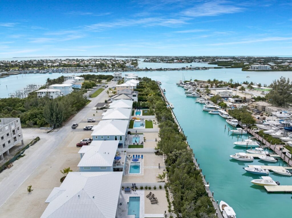 Aerial view of a waterfront neighborhood with modern houses, private pools, and boats docked along a turquoise canal. The scene includes lush greenery and a distant view of more houses and water under a blue sky.