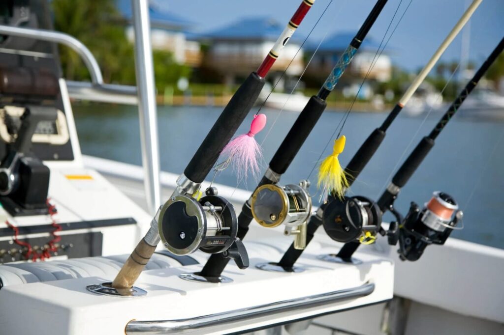 Four fishing rods with reels and colorful lures are lined up in holders on a boat, docked near water with blurred houses and trees in the background.