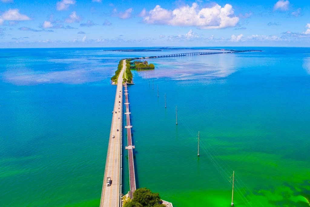 Aerial view of a long bridge stretching over clear turquoise water, connecting small islands under a blue sky with scattered clouds; cars drive along the bridge and power lines run parallel to the road.