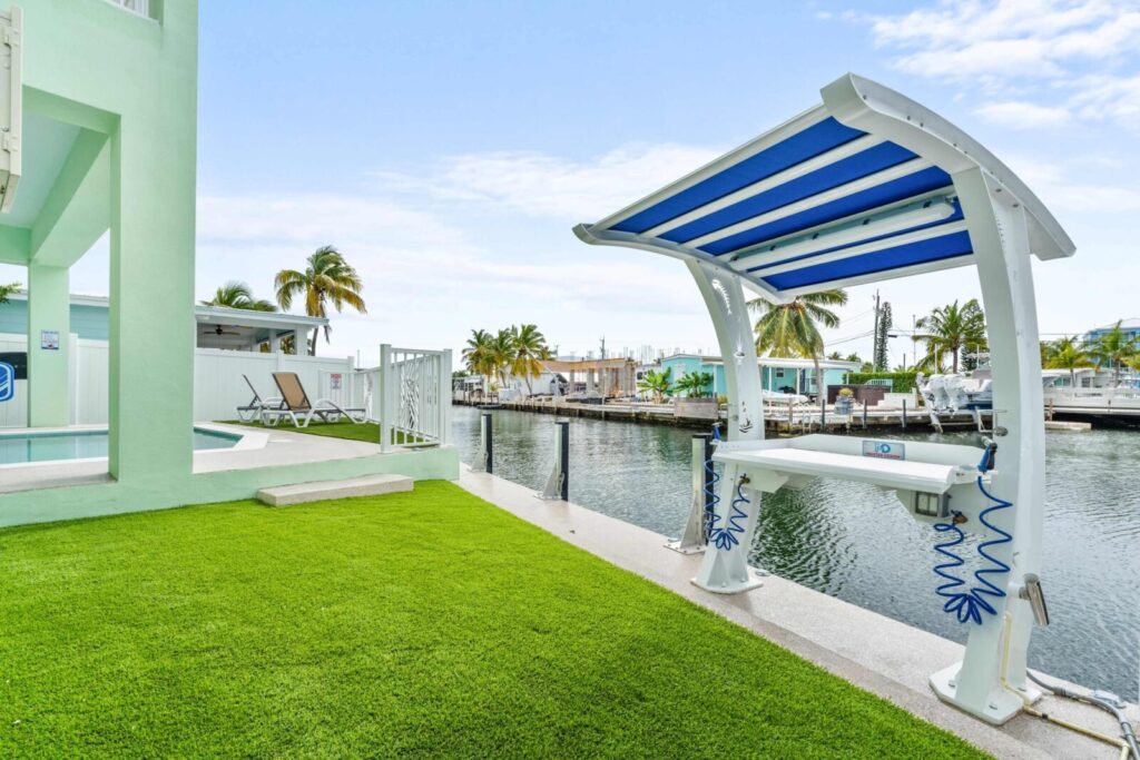 10 upgrades to increase your florida keys vacation rental revenue (2026) 4 A canal-side backyard with green artificial grass, a blue and white covered dockside fish cleaning station, and palm trees. Boats are docked along the canal under a partly cloudy sky.