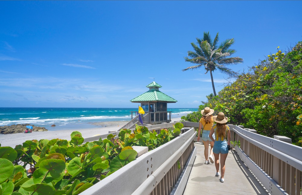 Two people in hats and summer outfits stroll along a wooden boardwalk toward a lifeguard tower on a sunny florida keys beach in february, surrounded by green vegetation, blue sky, and gentle ocean waves.