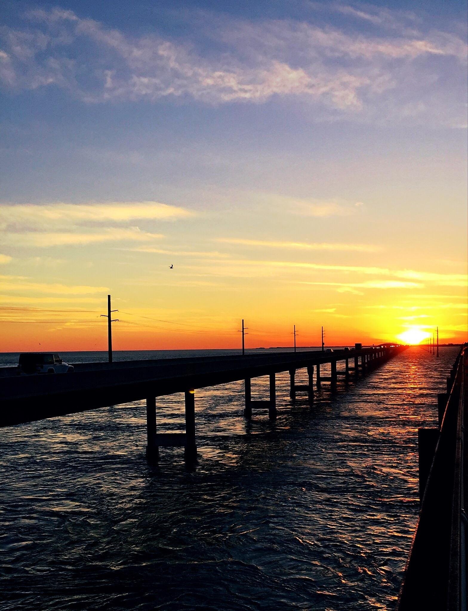 A long bridge stretches over the water at sunset in marathon fl, with golden sunlight reflecting on the waves. A car drives on the bridge, and power lines run alongside under a sky painted with orange, yellow, and blue hues.