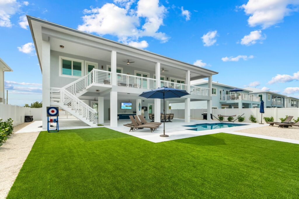 Modern two-story white house with a large balcony, outdoor lounge area, pool, and green lawn under a blue sky with scattered clouds. Patio chairs and umbrellas are set up around the pool.