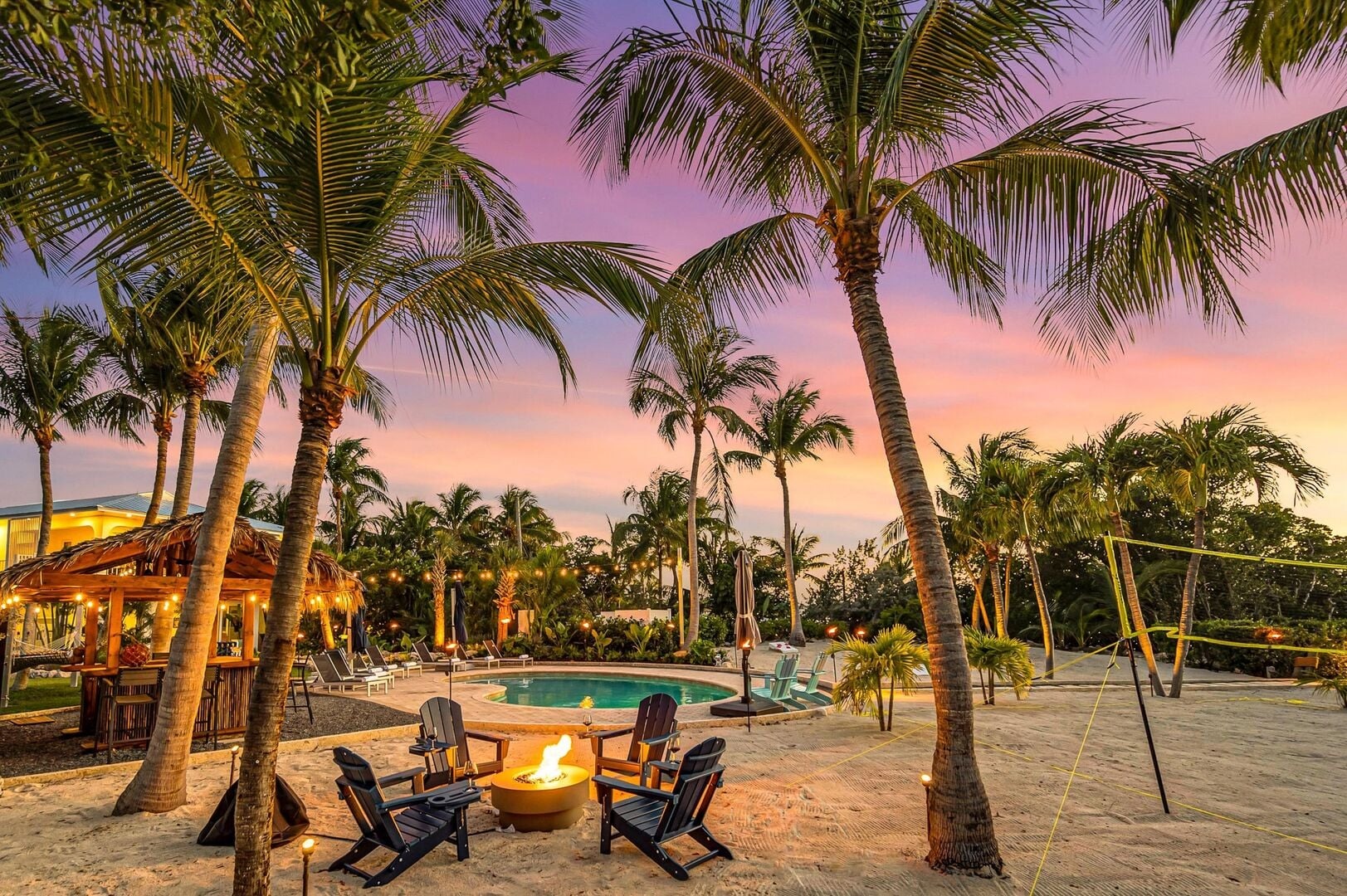 Tropical outdoor setting at sunset in marathon fl with palm trees, a fire pit surrounded by chairs, a pool, tiki hut, and a sandy area featuring a volleyball net. The sky glows in vibrant shades of purple and orange.