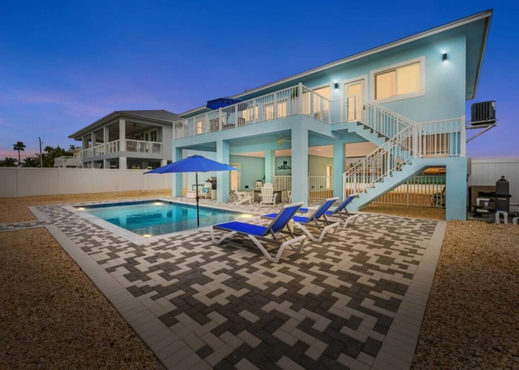 Two-story blue beach house with large windows, outdoor staircase, private swimming pool, poolside lounge chairs with blue umbrellas, and a patterned stone patio, all lit up at dusk.