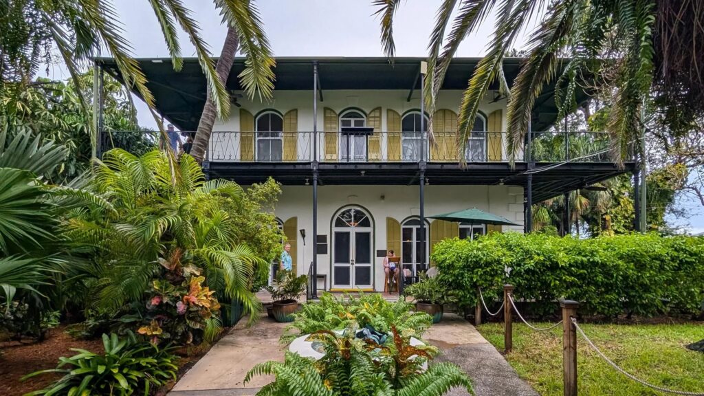 A two-story house with arched windows and doors, surrounded by lush tropical plants and palm trees, offers the perfect spring break in the florida keys. The upper floor features a black wrought-iron balcony and a pathway leading to the entrance.