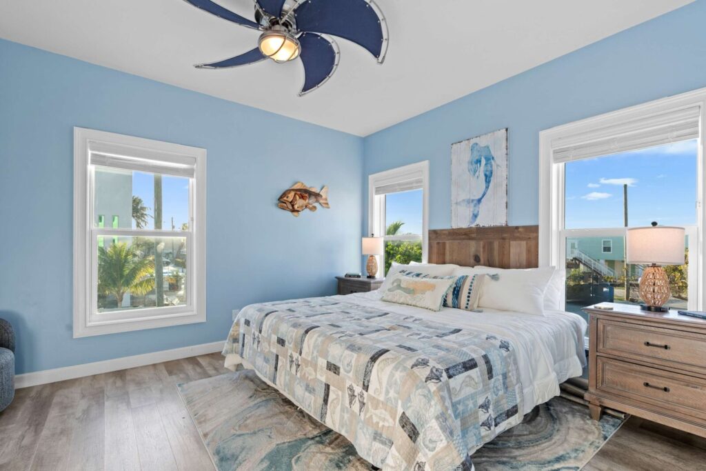 A bright bedroom with blue walls, a large bed with patterned bedding, wood nightstands with lamps, three windows letting in daylight, and beach-themed decor including a fish wall art and a ceiling fan shaped like flower petals.