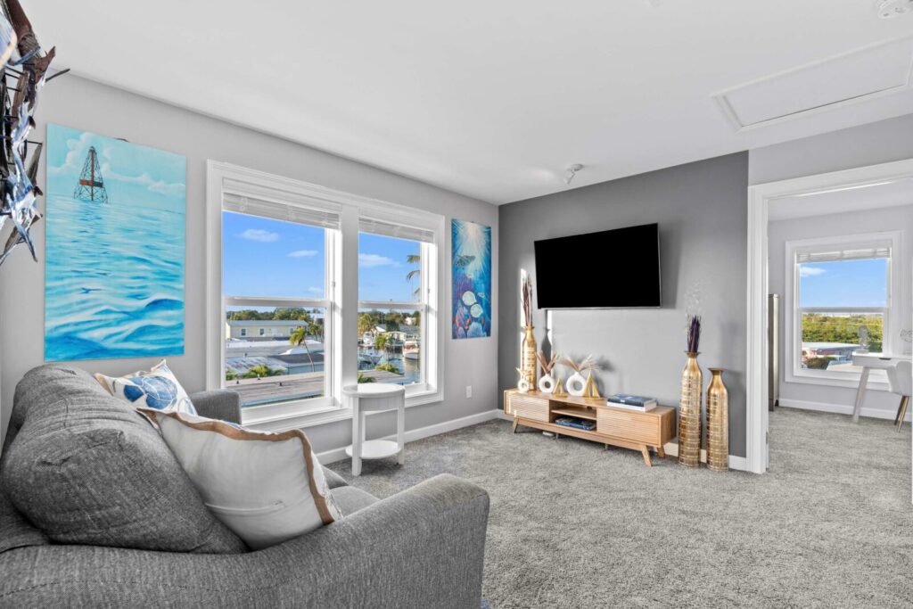 A bright living room with large windows, grey sofa, wall-mounted tv, coastal-themed decor, artwork of the sea, and decorative vases. An adjacent room with a desk is visible through an open doorway.