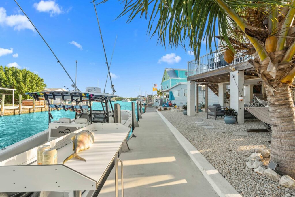 A dockside scene with fishing boats moored in turquoise water, a cutting table with a fish, a palm tree, and coastal houses with decks and outdoor seating under a clear blue sky.
