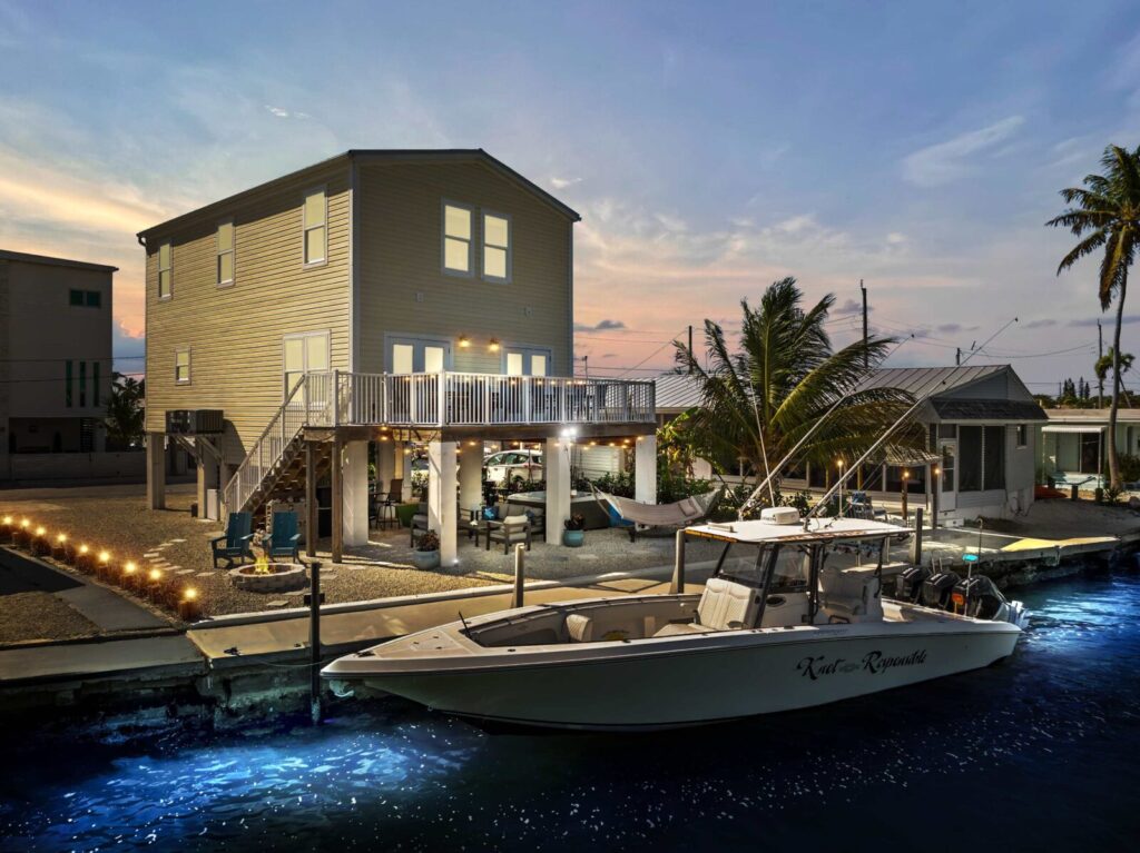 A modern, two-story waterfront house at dusk with glowing lights, a docked boat labeled reel or rejected, palm trees, and a hammock, creating a relaxed coastal atmosphere.