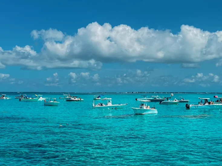 Boats float on clear turquoise water under a bright blue sky with scattered clouds. People snorkel and swim near the boats, enjoying the calm, tropical sea—perfect for a spring break in the florida keys.