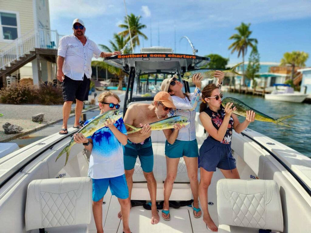 Four people stand on a boat holding freshly caught fish and pretending to kiss them, while a man stands at the back smiling. Palm trees and waterfront houses are visible in the background on a sunny day.