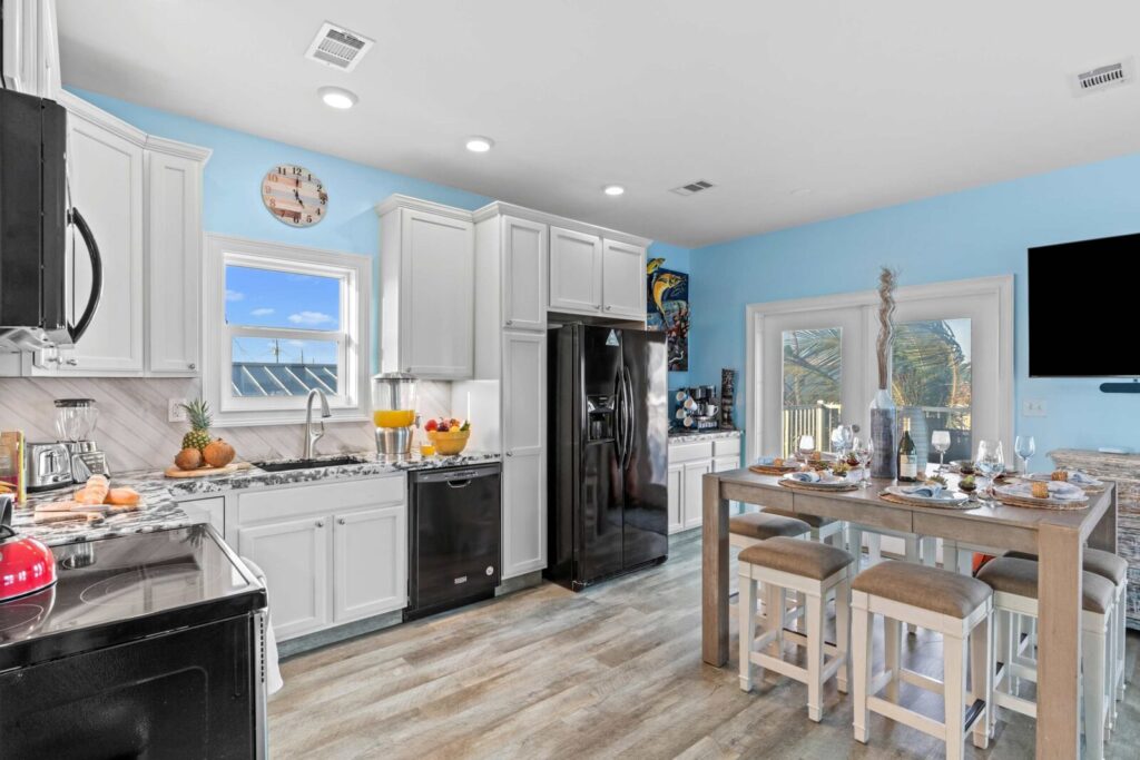 Bright, modern kitchen with blue walls, white cabinets, black appliances, and a breakfast bar set for four. Sunlight streams through windows, illuminating fruit on the counter and a beach view outside.