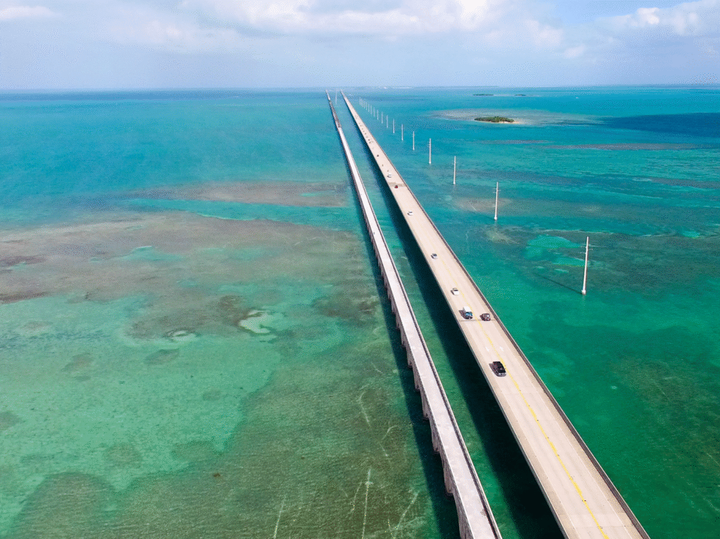 Aerial view of a long bridge with cars driving across turquoise ocean water, connecting small islands under a partly cloudy sky.