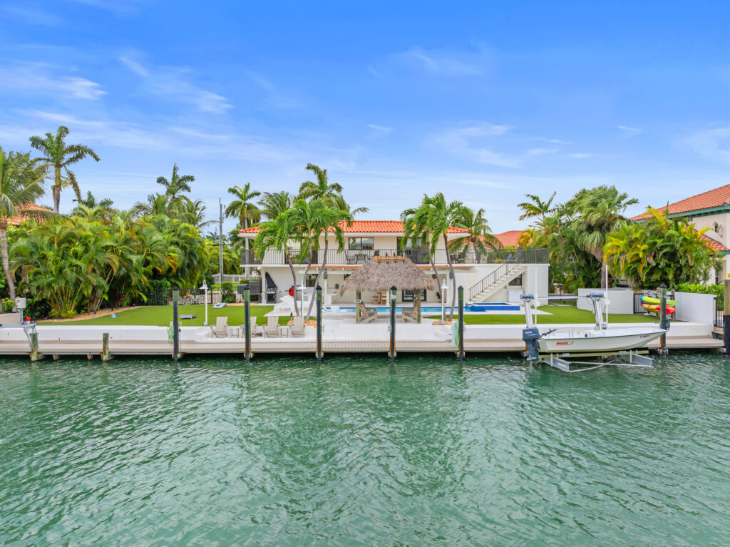 Waterfront property with a dock, tropical palm trees, lush landscaping, and a large house featuring a pool and outdoor seating area under a thatched roof, all overlooking calm, green water.