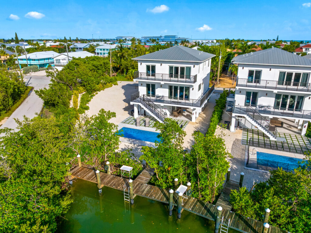 Isla sol: best waterfront vacation rental in marathon fl with pool & dock 15 Aerial view of two modern white houses with metal roofs, pools, and docks overlooking a canal, surrounded by greenery in a coastal neighborhood under a clear blue sky.