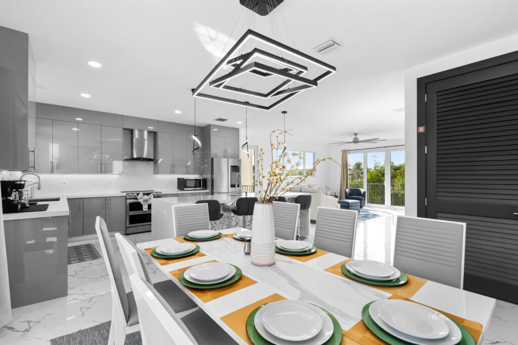 Isla sol: best waterfront vacation rental in marathon fl with pool & dock 4 Modern, open-plan kitchen and dining area with glossy gray cabinets, a white island, and a dining table set for six with yellow placemats and a vase of branches, leading into a bright living room with large windows.