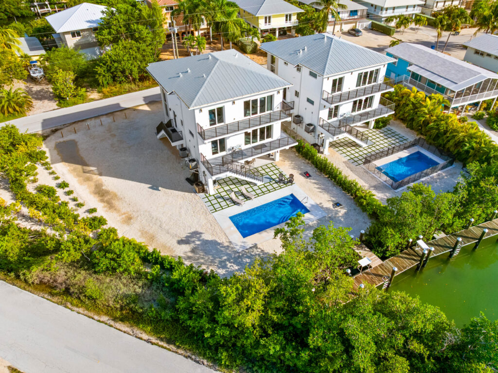 Isla sol: best waterfront vacation rental in marathon fl with pool & dock 11 Aerial view of two modern white houses with balconies, private pools, and sandy yards next to green trees, a dock, and calm water, surrounded by other residential homes.