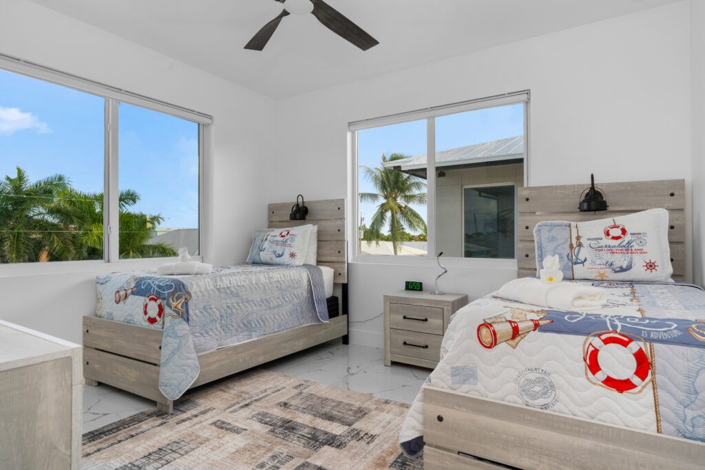 Isla sol: best waterfront vacation rental in marathon fl with pool & dock 7 Bright bedroom with two twin beds featuring nautical-themed bedding, a nightstand with an alarm clock between them, large windows showing palm trees, and a ceiling fan above.
