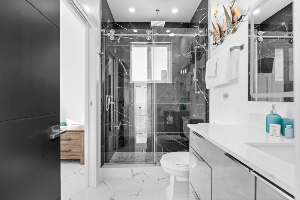 Isla sol: best waterfront vacation rental in marathon fl with pool & dock 8 Modern bathroom with black marble shower, glass door, white vanity, toilet, and marble floor. Blue and white accents on the sink, towels on the wall, and a decorative sailboat piece above the towel rack.