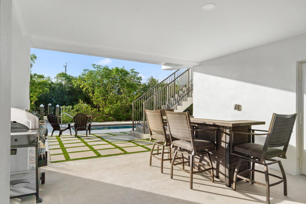 Isla sol: best waterfront vacation rental in marathon fl with pool & dock 10 Covered patio with a wicker dining set, stainless grill, and stairs leading up. Outside, a pool and fire pit area with adirondack chairs face a backdrop of green trees and a blue sky.