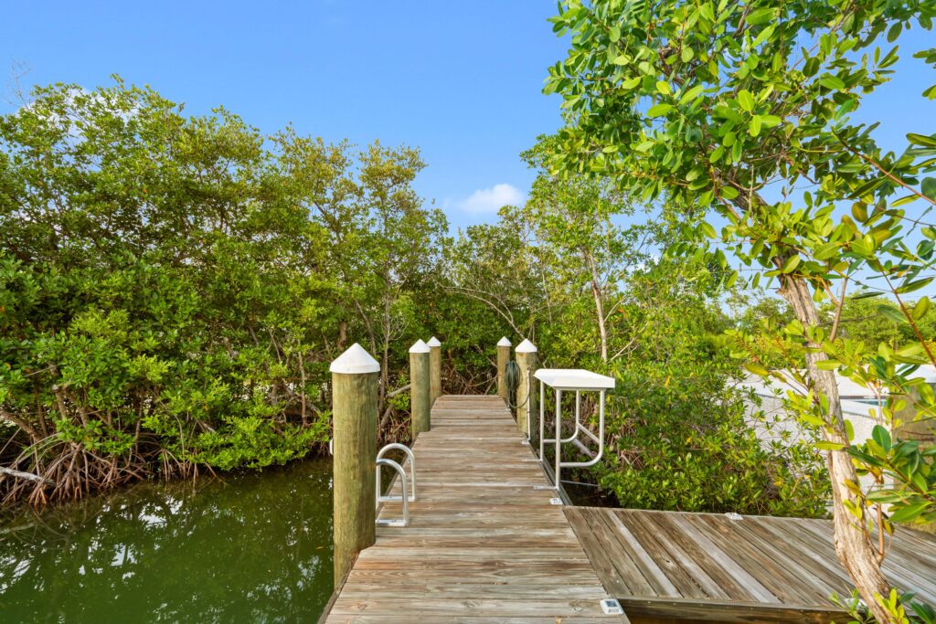 Isla sol: best waterfront vacation rental in marathon fl with pool & dock 12 A wooden dock extends over calm water, surrounded by lush green mangrove trees under a clear blue sky. White caps top the dock posts, and a metal ladder leads down to the water.