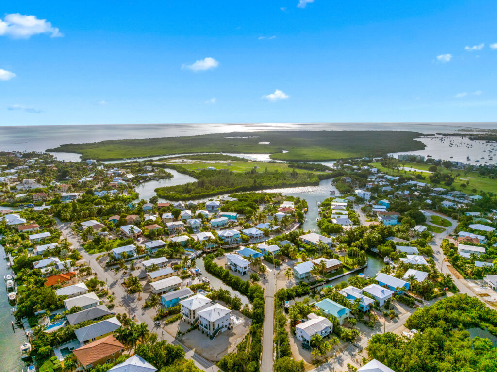 Aerial view of a coastal neighborhood with houses, winding roads, lush greenery, and water channels, bordered by a large expanse of water under a bright blue sky with a few clouds.