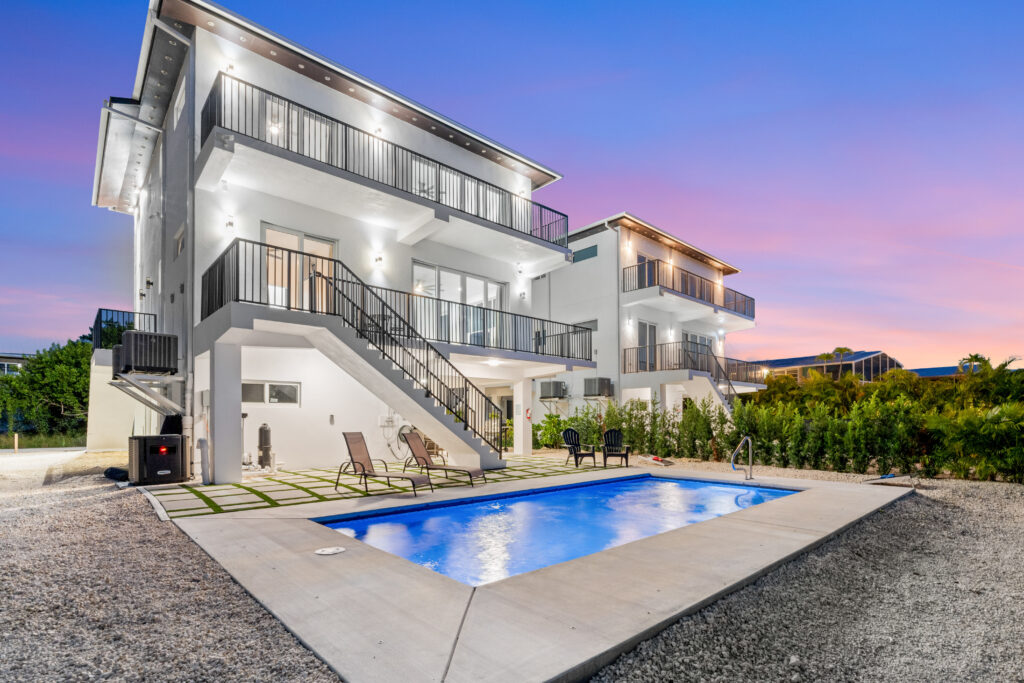 Isla sol: best waterfront vacation rental in marathon fl with pool & dock 2 Modern white two-story house with balconies and large windows, a small swimming pool in the backyard, lounge chairs, and landscaped surroundings during sunset with a colorful sky. Isla sol: waterfront vacation rental in marathon fl with pool & dock