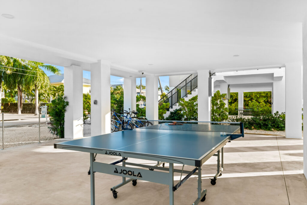 Isla sol: best waterfront vacation rental in marathon fl with pool & dock 6 A blue joola ping pong table sits in a bright, open-air covered area with white columns; bicycles and greenery are visible outside, along with a staircase and a sunny, tropical setting.