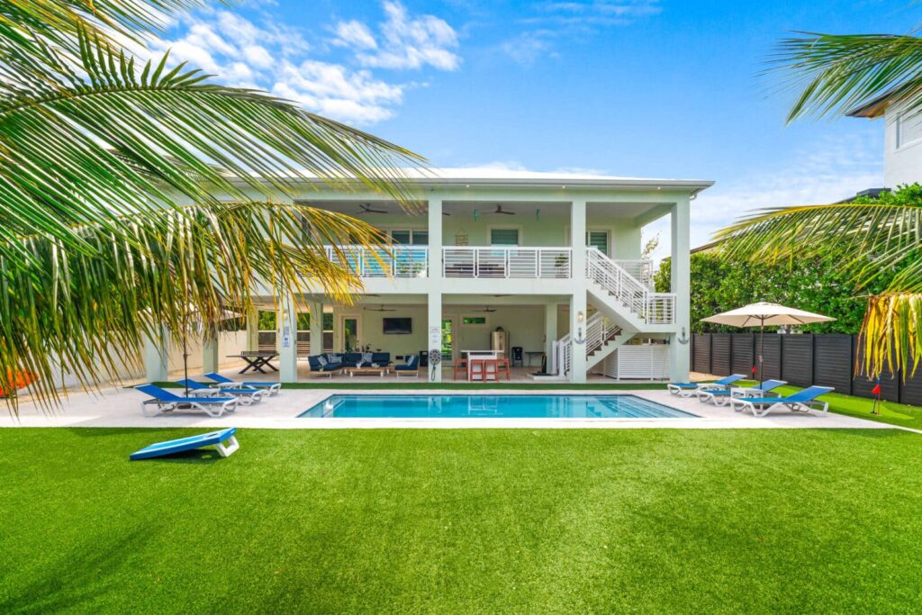 A two-story white modern house with a covered balcony, a central swimming pool, lounge chairs, and green artificial turf yard, framed by palm trees under a bright blue sky.