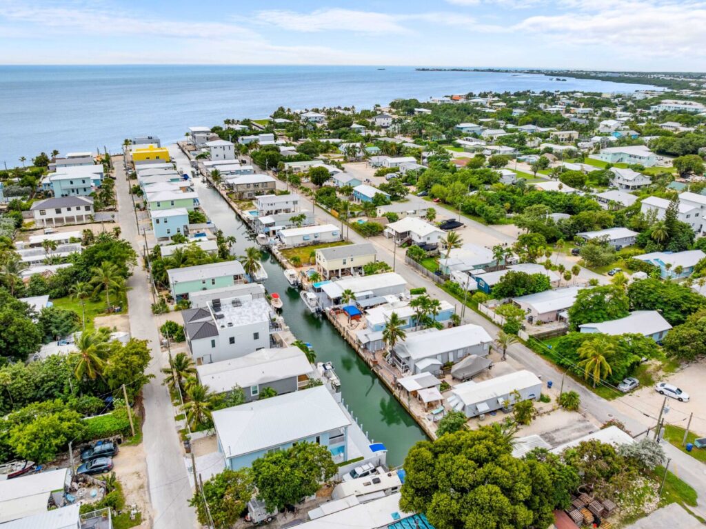 Aerial view of a coastal neighborhood with houses lining a narrow canal, lush greenery, palm trees, and the ocean visible in the background under a partly cloudy sky.