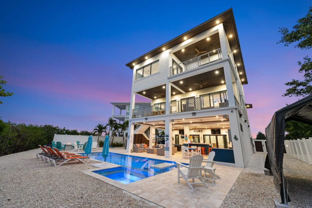 Modern three-story beach house with balconies, a pool, lounge chairs, outdoor dining area, and palm trees, set against a vibrant sunset sky with purple and pink hues.