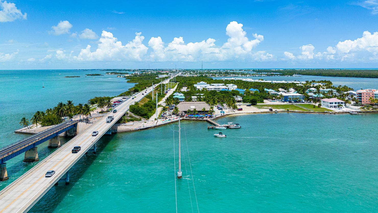 Aerial view of a bridge connecting islands over turquoise water, with cars crossing—showcasing getting around the florida keys—boats below, and lush trees, buildings, and clouds scattered across the bright blue sky.
