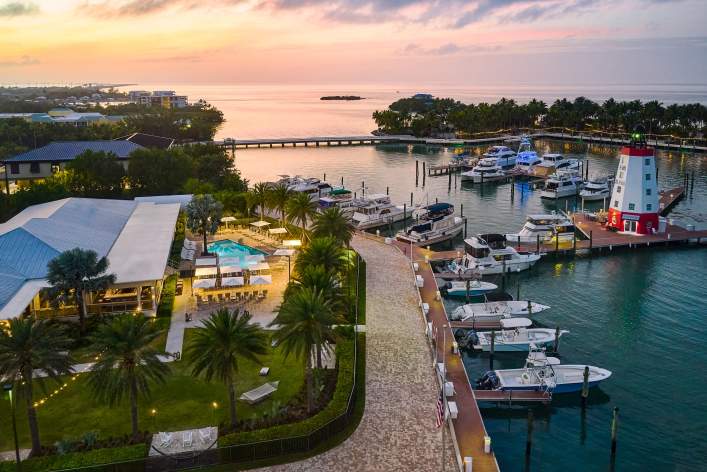 Aerial view of a marina at sunset, with boats docked in the harbor, a red and white lighthouse, palm trees, a swimming pool, and nearby buildings surrounded by lush greenery and calm water.